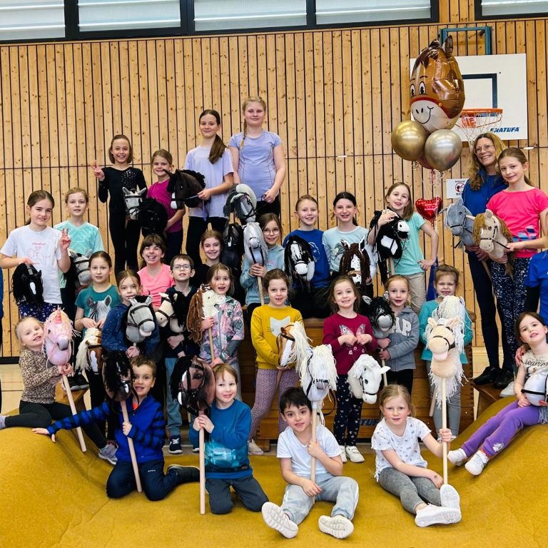 Eine Gruppe von Kindern mit Holzpferden und Ballons in einer Turnhalle.
