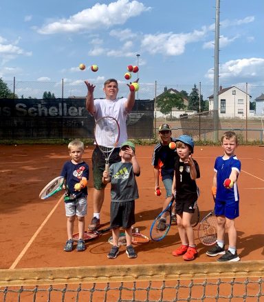 Eine Gruppe von Kindern und einem Trainer auf einem Tennisplatz mit Bällen und Schlägern.