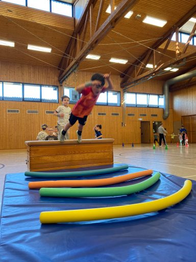 Ein Kind springt über eine Holzbank, während andere Kindern im Hintergrund spielen.