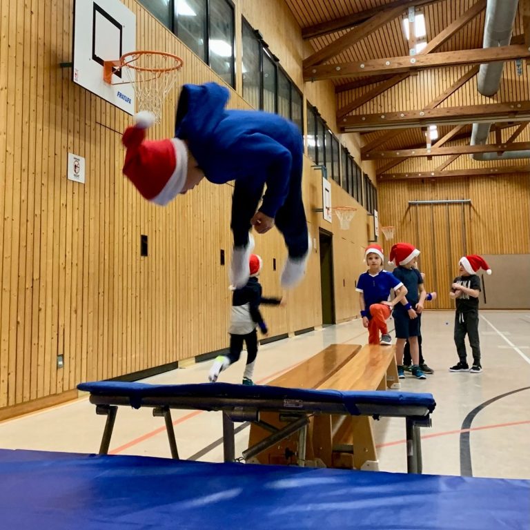 Junge springt mit Weihnachtsmütze über ein Trampolin auf eine Matte in einer Sporthalle.