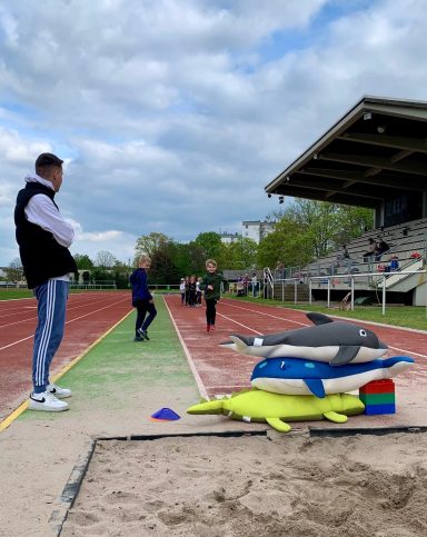 Athleten trainieren auf einer Leichtathletikbahn, Spielgeräte liegen daneben.