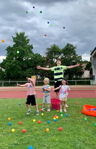 Kinder spielen im Freien mit bunten Bällen und lachen unter einem bewölkten Himmel.