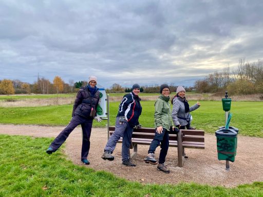 Vier Personen posieren humorvoll auf einem Weg im Freien, mit einer Bank im Hintergrund.