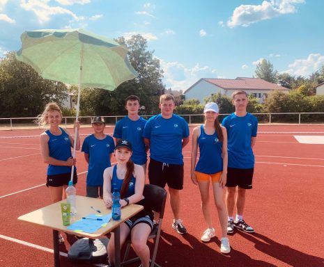 Grupp von sieben Personen in blauer Sportkleidung auf einem Tennisplatz, unter einem Sonnenschirm.